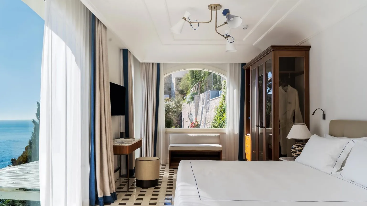 Geniş, aydınlık ve lüks bir otel yatak odasında, sol tarafta göz alabildiğince uzanan masmavi denizin huzur veren panoramik manzarası, odanın ortasındaki kemerli pencereden görünen yeşil bahçe ve taş duvarlarla çevrili bir avlu ile birleşerek unutulmaz bir tatil atmosferi yaratıyor.
