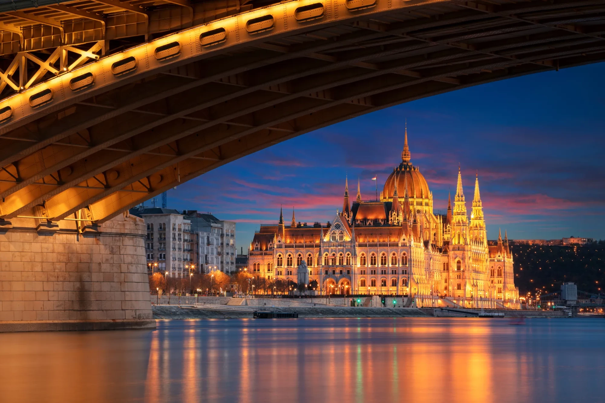 Tuna Nehri üzerinde, Margaret Köprüsü'nün altından çekilmiş bu büyüleyici Budapeşte fotoğrafında, altın rengi ışıklarla aydınlatılmış, görkemli kubbesi ve kuleleriyle Macaristan Parlamento Binası, alacakaranlıkta gökyüzünün ve suyun yansımalarıyla parlıyor.