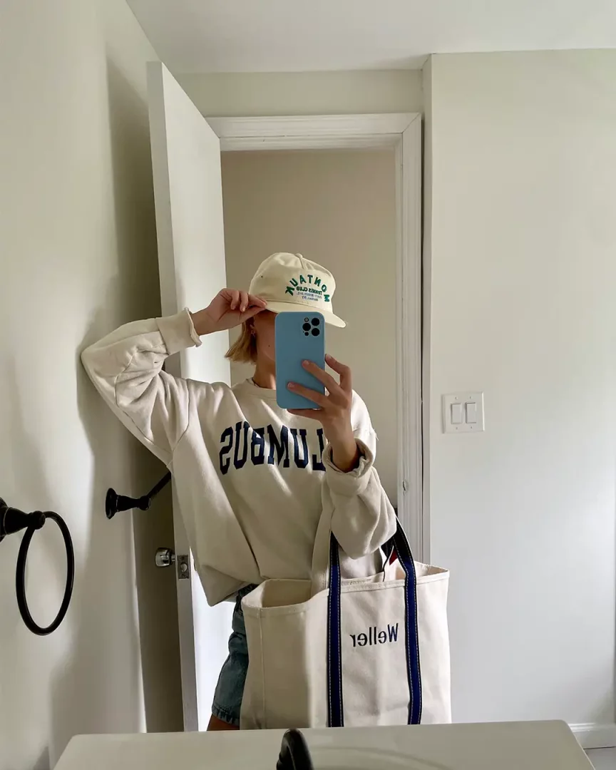 Ayna karşısında krem rengi sweatshirt ve şapka giymiş, yüzünü mavi telefonuyla kapatan bir kişi, lacivert şeritli ve Weller yazılı büyük boy kanvas omuz çantasını yanında tutarak günlük stilini tamamlıyor.