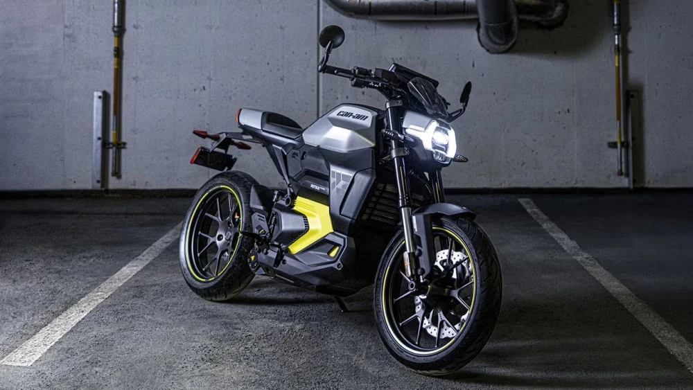 Gümüş gri ve siyah renklerin hakim olduğu, neon sarı detaylı jantlara ve yan panellere sahip, farları yanan modern tasarımlı Can-Am Pulse elektrikli motosiklet, loş ışıklı bir otoparkın beton zemini üzerinde park etmiş halde duruyor.