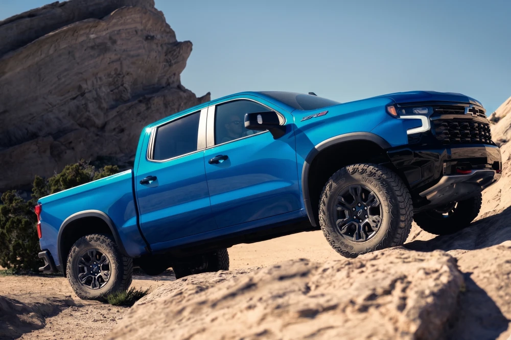 Mavi Chevrolet Silverado ZR2 pickup arazi aracı, engebeli ve kayalık bir off-road arazide eğimli bir şekilde durarak off-road yeteneğini sergiliyor; geniş yanaklı lastikleri ve yükseltilmiş şasisi bu zorlu koşullara uygunluğunu vurguluyor.