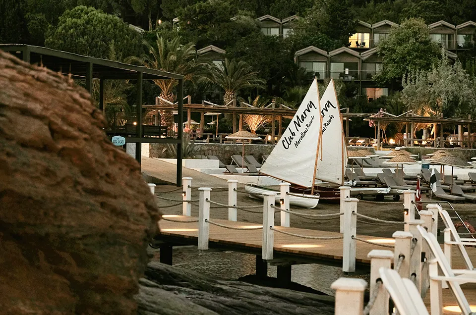 Soldaki büyük kayanın arkasından görünen Club Marvy Marvellous Resorts'un akşamüstü ışıklarıyla aydınlatılmış plajı, ahşap iskelesi, iki yelkenli teknesi, güneşlenme şezlongları ve palmiye ağaçları arasındaki konaklama birimleriyle sakin bir tatil cenneti sunuyor.