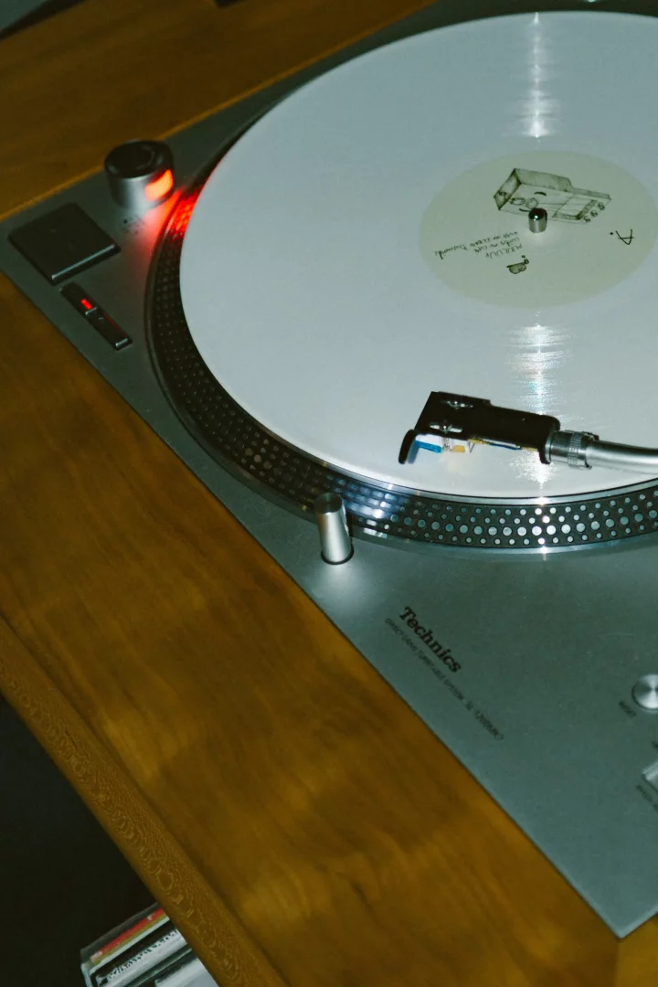 Üzerinde beyaz bir plağın bulunduğu, açma düğmesindeki kırmızı ışığıyla Technics SL-1200MK model bir pikabın ahşap bir yüzeyde konumlandırılmış hali yakın çekim olarak gösterilirken, plağın yüzeyine hassasça inmiş iğnesiyle detaylı bir dinleme deneyimine işaret ediyor.