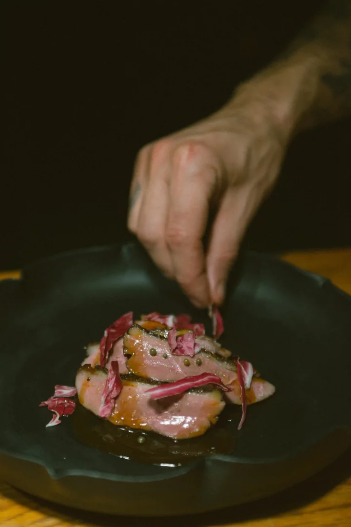 Koyu renkli bir tabağa özenle yerleştirilmiş az pişmiş et dilimleri ve zengin sosun üzerine, bir şefin eli mor renkli radicchio ve kaparilerle Amsterdam'ın sofistike mutfak anlayışını yansıtan son dokunuşları yapıyor.