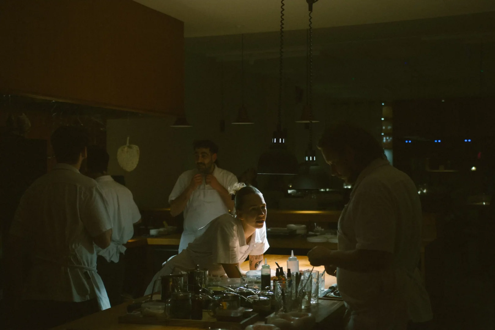 Loş ışıklı bir restoran mutfağında, tezgah başında çalışan genç bir kadın şef ve etrafındaki diğer şefler, modern asma lambaların aydınlattığı yoğun ama işbirlikçi bir atmosferde yemek hazırlığı yapıyor, özenli ve profesyonel bir çalışma ortamını yansıtıyor.