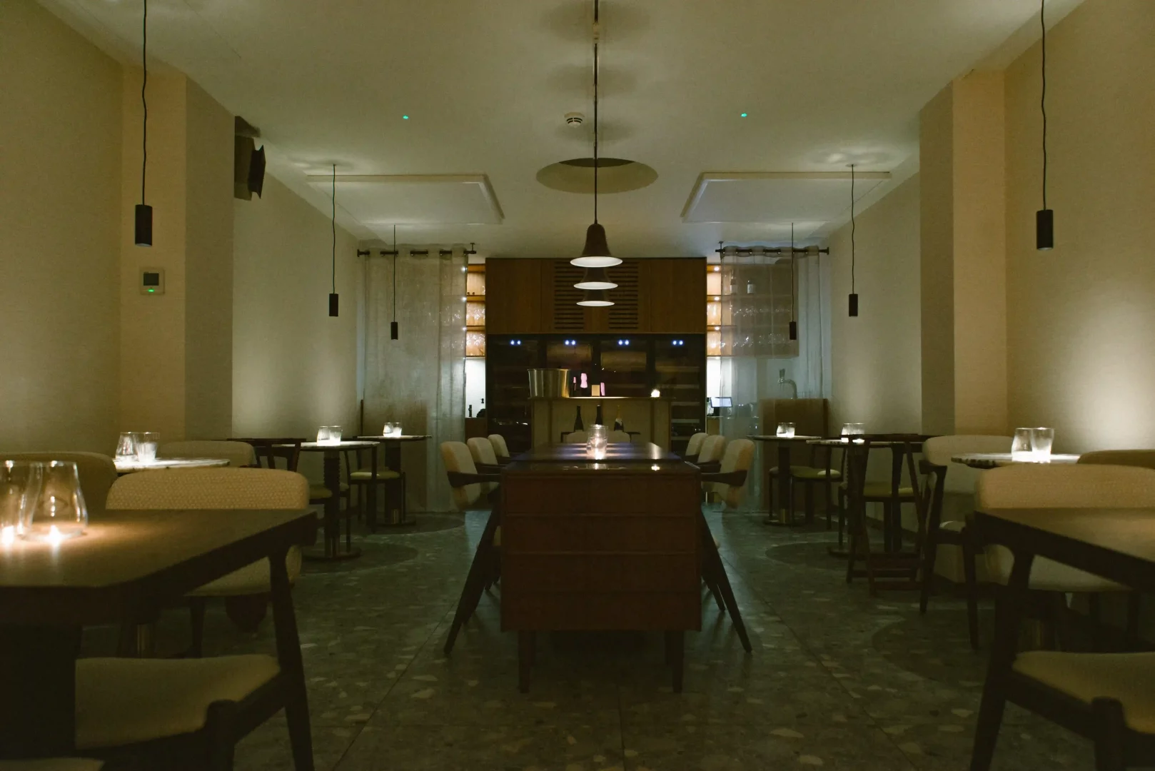 Loş ışıklı, modern tasarımlı bir restoranda, ortada uzun bir ahşap masa ve etrafında krem renkli sandalyelerle, duvar kenarlarında şık küçük masaların mum ışıklarıyla aydınlatıldığı, davetkar ve sakin bir yemek ortamı.