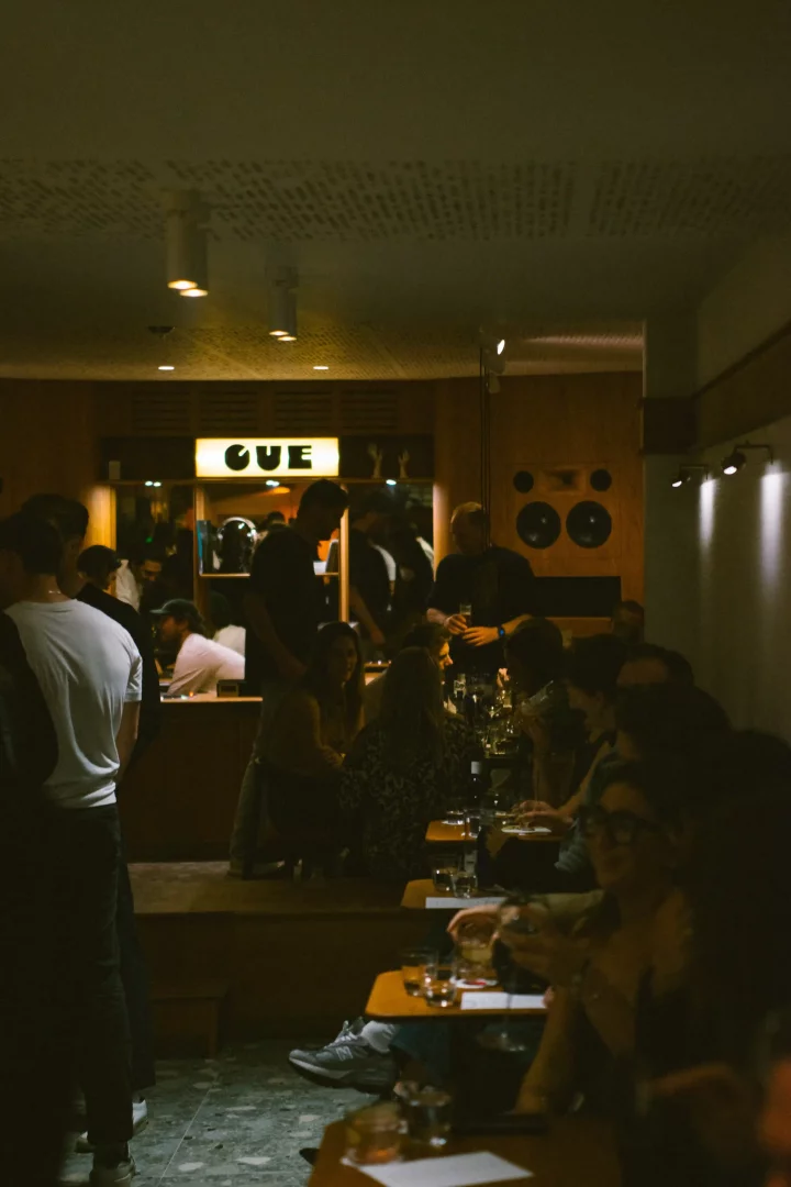Cue adlı mekanın loş ışıklı, ahşap detaylarla dolu iç mekanında, parlayan CUE tabelasının altında ayakta ve masalarda oturan çok sayıda insan içkileriyle sohbet ediyor, duvardaki hoparlörler eşliğinde canlı bir atmosferde sosyalleşiyor.