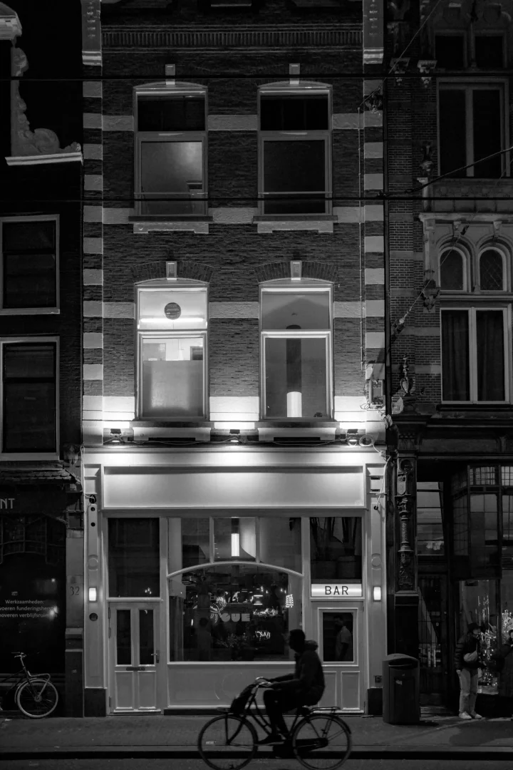 Amsterdam'da geceleri tuğla cepheli, ışıklarla aydınlatılmış bir barın önünden bisikletle geçen bir siluet, şehrin hareketli atmosferini ve gece hayatını yansıtan dinamik bir şehir sahnesi sunuyor.
