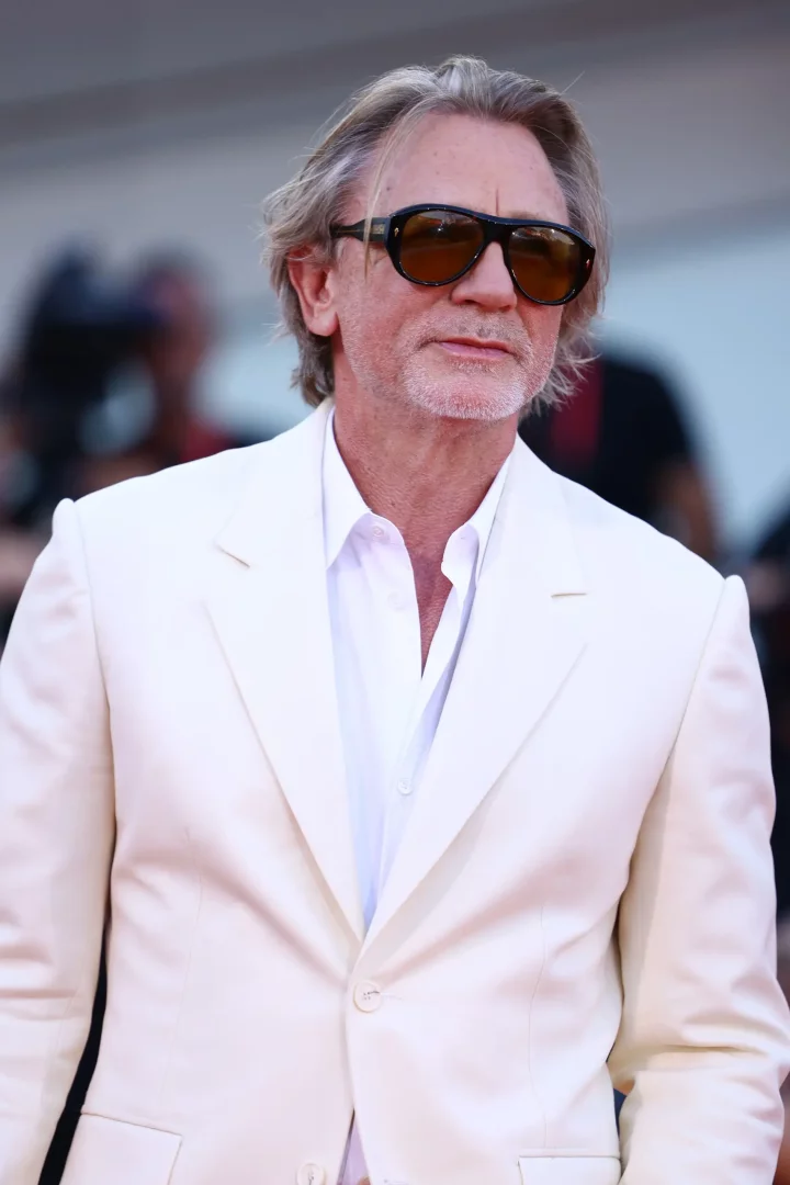 Krem rengi bir takım elbise ceketi ve açık yakalı beyaz bir gömlek giymiş, koyu renk güneş gözlükleri, gri-sarışın saçları ve açık gri sakallarıyla Daniel Craig, bulanık bir kırmızı halı etkinliği arka planında hafifçe sola dönük poz veriyor.