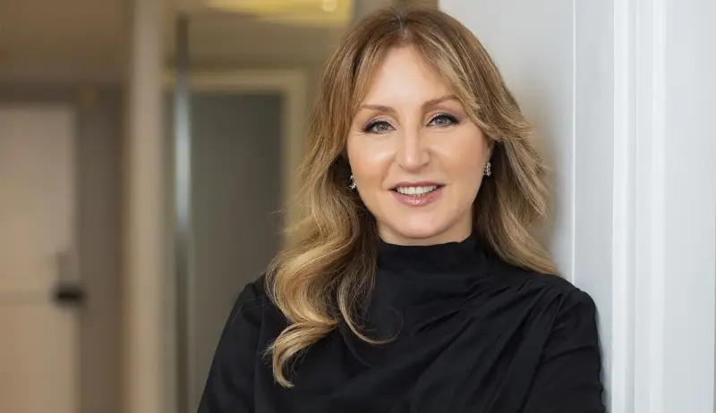 Dermatoloji Uzmanı Betül Şengör, açık sarı dalgalı saçları, zarif siyah boğazlı bluzu ve şık küpeleriyle, hafif makyajı ile kameraya samimi bir gülümsemeyle bakarak, dermatoloji konusundaki uzmanlığını ve sıcak kişiliğini vurguluyor.