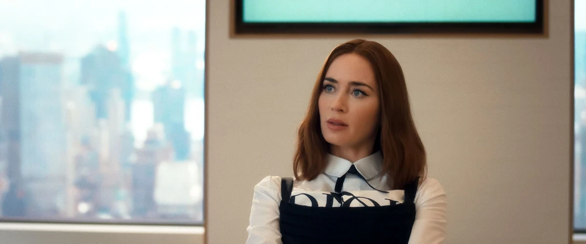 Şeytan Marka Giyer 2 filminde Emily Blunt tarafından canlandırılan Emily Charlton, beyaz yakalı gömleğinin üzerine giydiği ve moda dünyasında markaların artan gücünü simgeleyen belirgin bir logoya sahip siyah giysisiyle, New York siluetini yansıtan modern ofis penceresi önünde, dijitalleşen Runway evreninin yeni kurallarını değerlendiren düşünceli bir ifadeyle duruyor.