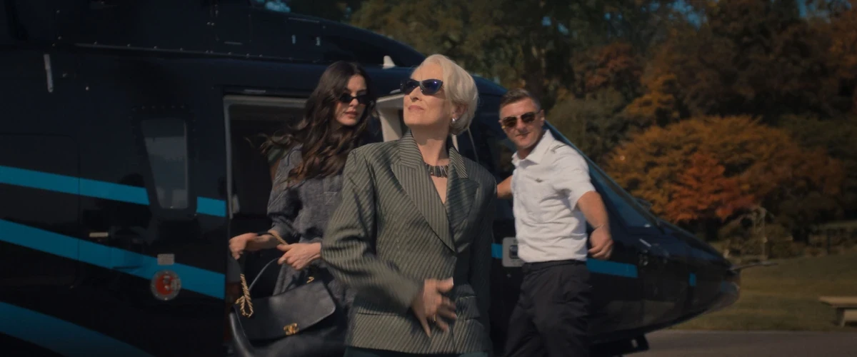 Meryl Streep'in canlandırdığı Miranda Priestly, çizgili blazerı ve güneş gözlükleriyle siyah bir helikopterden karizmatik bir şekilde inerken, Anne Hathaway'in canlandırdığı Andy Sachs hemen arkasında elinde çantasıyla ona eşlik ediyor ve bir pilot da kapıda beliriyor; arka planda sonbahar yapraklarıyla dolu ağaçlar görülüyor.