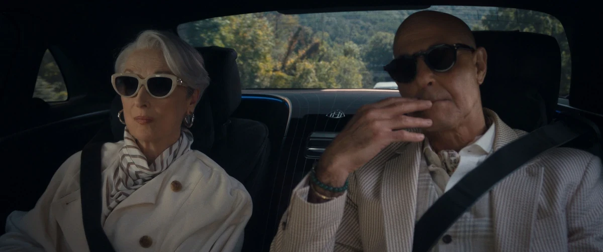 The Devil Wears Prada 2 filminden Miranda Priestly ve Nigel, şık güneş gözlükleri ve kıyafetleriyle lüks bir arabanın arka koltuğunda seyahat ederken görülüyor; Miranda solda, hafifçe sağa dönük, sakin bir ifadeyle dururken, Nigel ise sağda, elini ağzına yakın tutarak düşünceli bir anı yansıtıyor, bu dinamik ikilinin karizmatik araba sahnesi filmin ruhunu yansıtıyor.