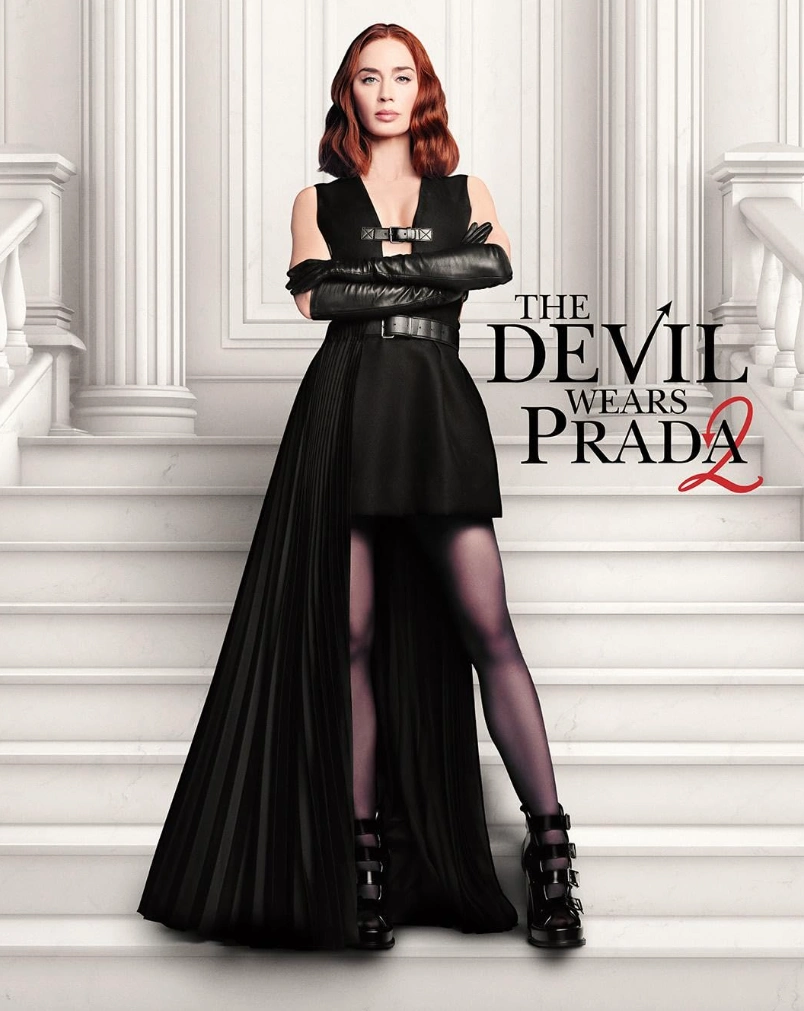 Emily Blunt, Emily Charlton rolünde, The Devil Wears Prada 2 filminin iddialı kostümleri arasında öne çıkan, korse etkili siyah bir üstü kısa, arkası pilili uzun eteği olan elbiseyi, uzun deri eldivenleri ve tokalı botları giyerek beyaz bir merdivende kolları bağlı, meydan okuyan bir ifadeyle duruyor.