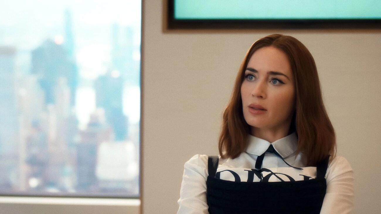 Emily Blunt, Şeytan Marka Giyer filmindeki Emily Charlton karakteriyle, beyaz yakalı gömleği üzerine giydiği siyah askılı ve önünde belirgin DIOR yazısı olan modern kostümüyle, soluk mavi şehir siluetinin göründüğü pencerenin önünde hafifçe sağa dönük, düşünceli bir ifadeyle duruyor.