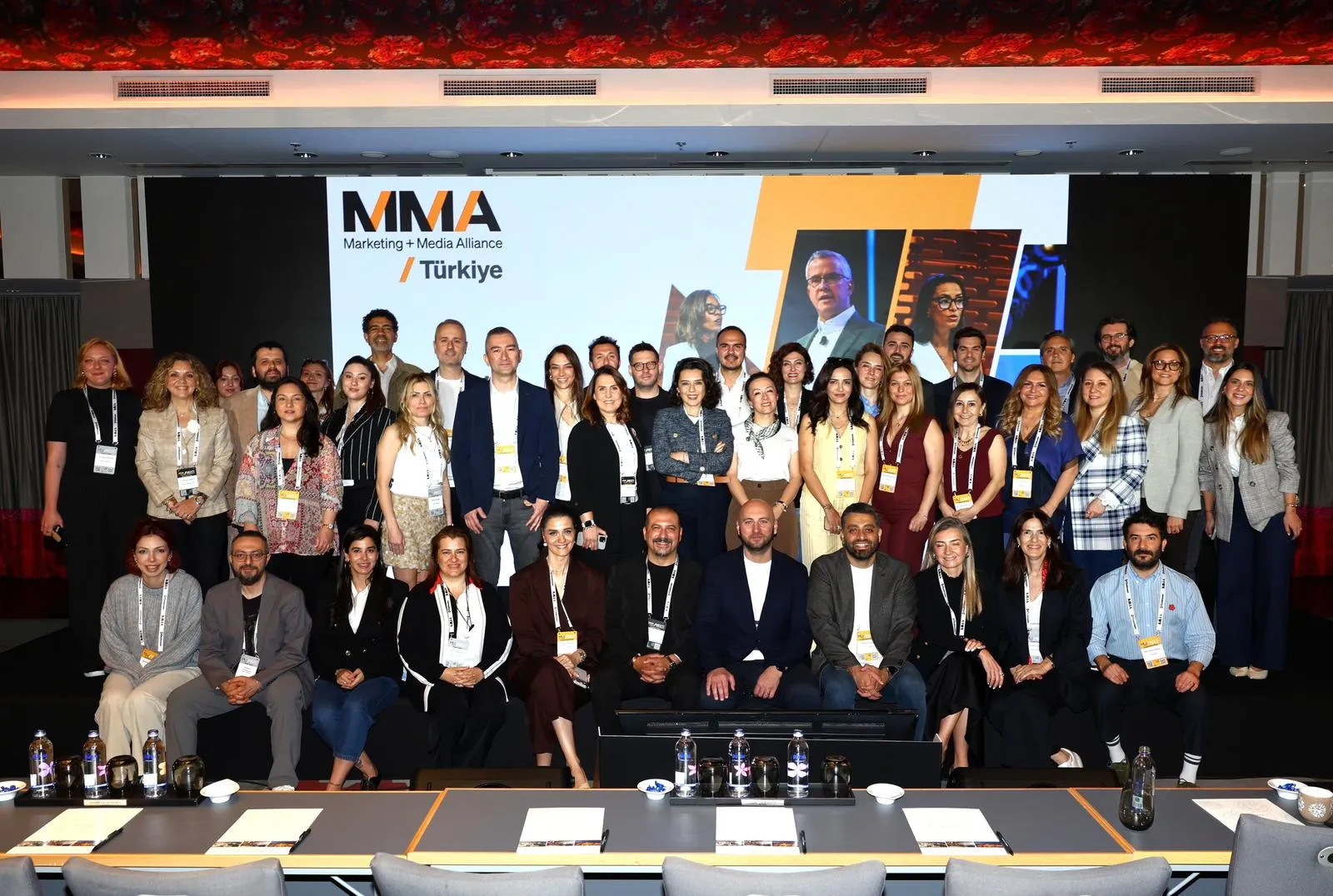 MMA Marketing + Medya İttifakı Türkiye logosunun büyük bir ekranda belirgin şekilde görüldüğü bir konferans salonunda, farklı yaş ve cinsiyetten pazarlama ve medya profesyonelleri, etkinliğin ruhunu yansıtan kalabalık bir grup fotoğrafı için bir araya gelmiş.