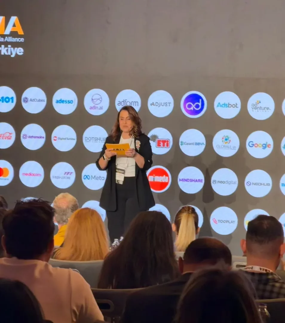Sahne üzerinde, arkasında birçok şirketin logosu ve MMA Türkiye yazısı bulunan bir fonda, Didem Namver siyah blazer ceketi ve beyaz bluzuyla elinde sarı not kağıtları tutarak dikkatle sunumunu yapıyor, ön planda ise dinleyicilerin başları görülüyor.