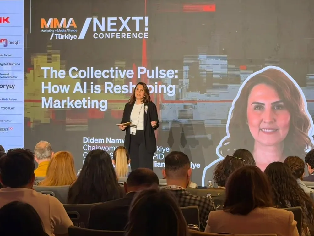 MMA NEXT! Konferansı'nda sahnedeki Didem Namver, büyük bir ekranda Yapay Zeka'nın Pazarlama'yı nasıl yeniden şekillendirdiğini gösteren sunumu yaparken izleyicilere dönük konuşuyor ve ekranda kendisinin gülümseyen bir portresi de görülüyor.