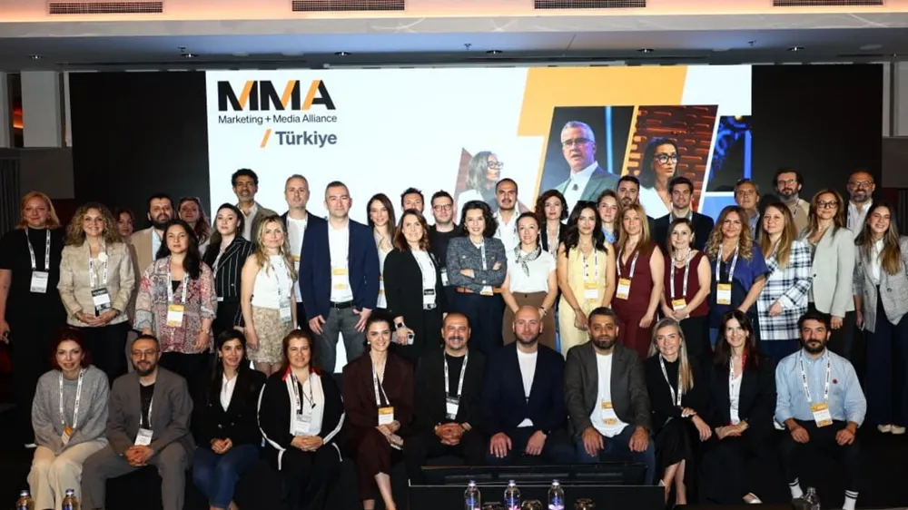MMA Marketing + Media Alliance Türkiye'nin çeşitli kademelerinden yöneticiler ve profesyonellerin, markanın logosunun ve geçmiş etkinlik görsellerinin yer aldığı büyük bir ekran önünde topluca poz verdiği, katılımcıların yaka kartlarıyla kurumsal kimliği yansıttığı canlı bir etkinlik fotoğrafı.