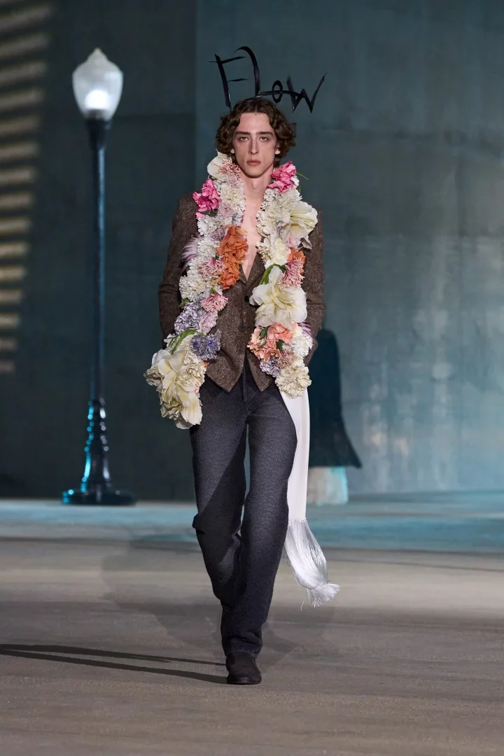 Dior Cruise 2027 koleksiyonundan bir erkek model, Los Angeles podyumunda kahverengi, açık önlü ceketinin üzerinde pembe, beyaz, mor ve krem tonlarında devasa bir çiçek çelengi ve sol tarafında sarkan beyaz püsküllü bir şal ile yürüyor; bu görünüm, Hollywood ve California poppy çiçeklerinden alınan ilhamı modern bir yorumla sunuyor.