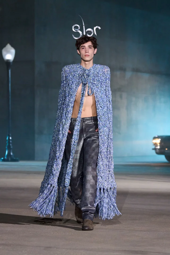 Jonathan Anderson'ın Dior için tasarladığı ilk cruise koleksiyonu olan Dior Cruise 2027'nin Los Angeles defilesinde, göğsü açık bir erkek model, uzun, püsküllü, mavi tonlarda örgü pelerin ve parlak, kareli desenli pantolonla yürüyor.