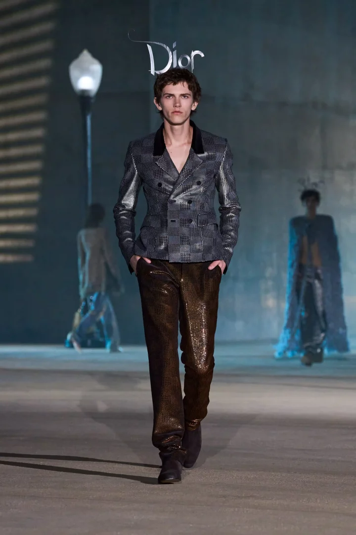Podyumda yürüyen genç erkek model, Jonathan Anderson'ın Dior Cruise 2027 koleksiyonundan parlak, ekose desenli çift düğmeli ceket ve ışıltılı kahverengi pantolonuyla Hollywood Estetiği'ni modern bir yorumla sunuyor; arkada hafifçe görünen figürler ve Dior logosu defilenin teatral atmosferini pekiştiriyor.
