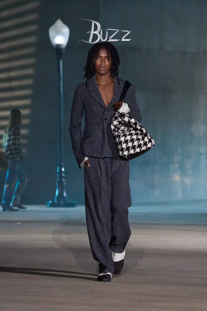 Rastalı saçlı genç bir erkek model, Jonathan Anderson'ın Dior Cruise 2027 koleksiyonundan koyu gri, hafif ışıltılı, bol kesim bir takımla podyumda yürürken, kolunda siyah beyaz kazayağı desenli bir çanta taşıyor ve arka planda loş bir sokak lambası ile havada asılı duran Buzz yazısı Hollywood atmosferini vurguluyor.