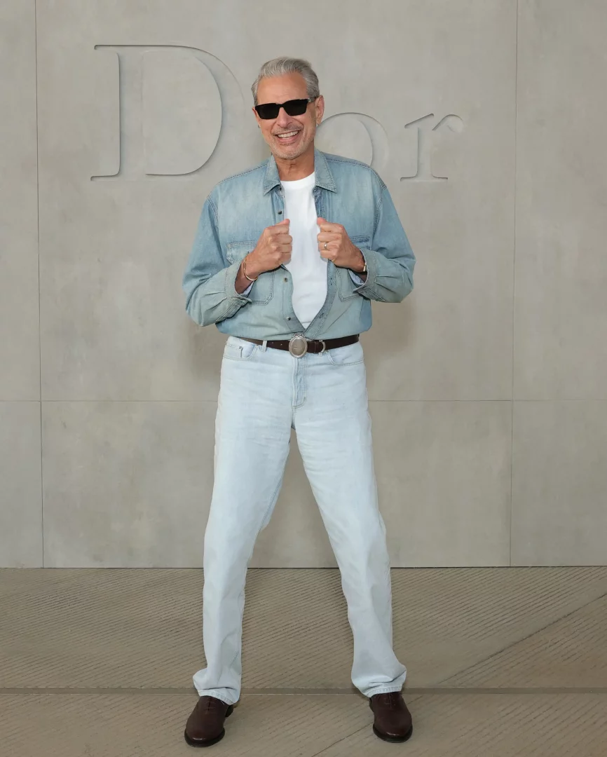 Dior Cruise 2027 defilesinin dikkat çeken konuklarından Jeff Goldblum, üzerinde açık renk bir kot gömlek, beyaz tişört, kot pantolon ve güneş gözlüğüyle, gömleğinin yakalarını tutarak neşeli bir ifadeyle objektife gülümsüyor, arkasında ise büyük Dior logosu bulunuyor.