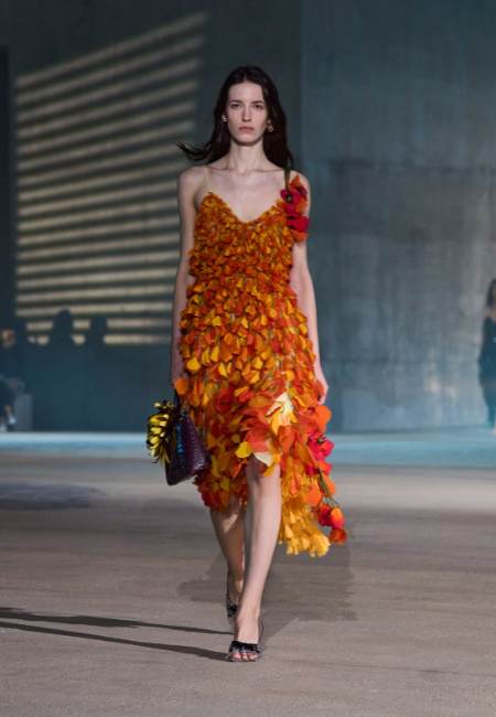 Dior Cruise 2027 koleksiyonundan bir manken, Kaliforniya gelinciklerini anımsatan, turuncu ve sarı yapraklardan oluşan katmanlı, sol omuzunda kırmızı çiçek detayları bulunan bir elbiseyle podyumda yürüyor, koleksiyonun doğal ve romantik estetiğini vurguluyor.