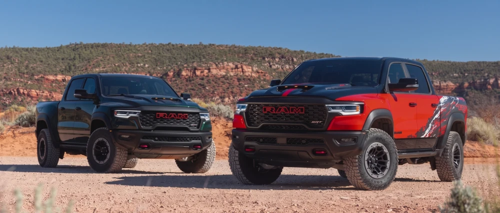 Çöl manzarasının ortasında, toprak bir yolda duran, sağ tarafta kaputu siyah detaylı ve yanları dikkat çekici kırmızı-siyah grafik desenli bir Ram 1500 TRX pickup ile sol tarafta koyu yeşil renkte başka bir güçlü Ram TRX pickup aracı.