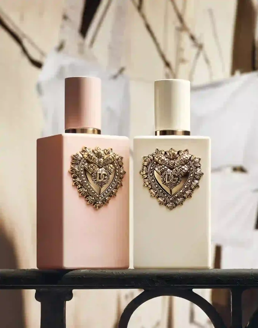 Dolce & Gabbana'nın My Devotion Eau de Parfum koleksiyonundan, zarif altın kalp ve iç içe geçmiş DG harfleriyle özenle süslenmiş, biri pastel pembe diğeri krem rengi olmak üzere iki şık parfüm şişesi, koyu ferforje bir korkuluğun üzerinde sergileniyor.