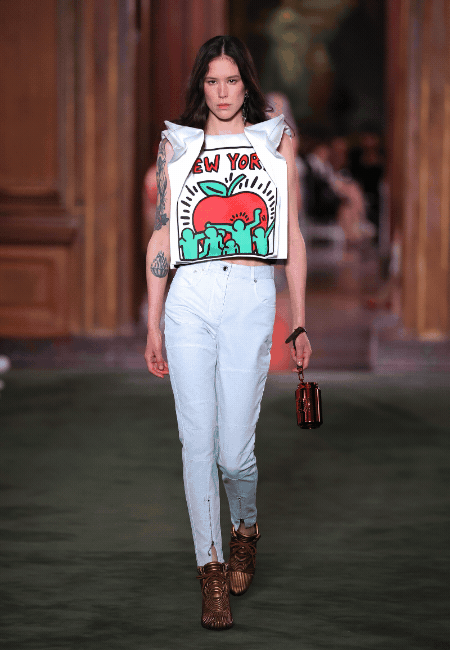 Louis Vuitton Cruise 2027 defilesinden, omuzları yapılı beyaz Keith Haring temalı New York elmalı ve dans eden figürlü crop top, açık renk pantolon ve metalik kahverengi çanta ile bronz botlar giyen bir model, The Frick Collection'ın zarif salonlarında podyumda yürüyor.
