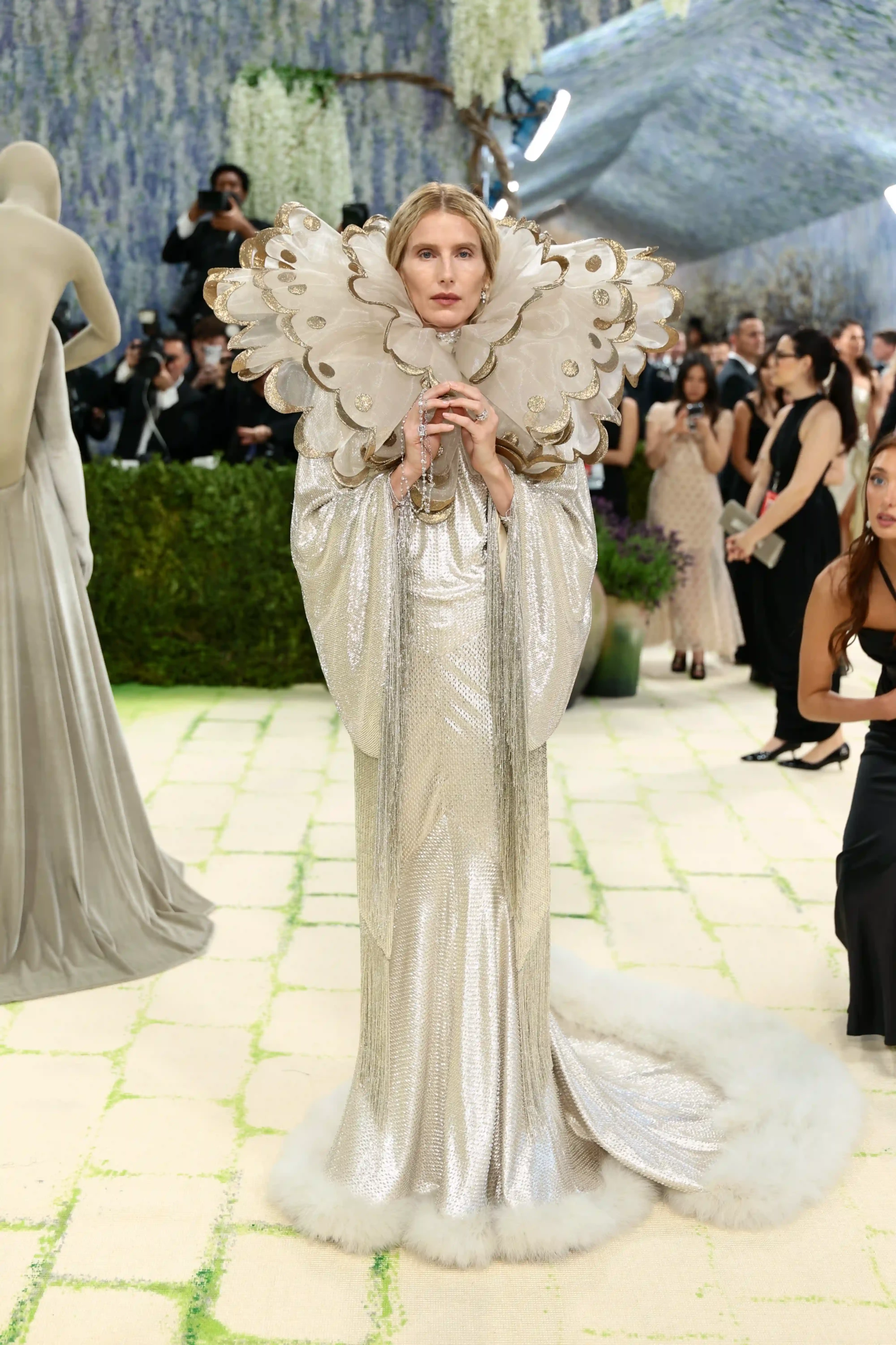Dree Hemingway, 2026 Met Gala kırmızı halısında, gümüş rengi payetlerle işli, uzun, etek ve kol uçları beyaz tüylü bir elbise içinde, yüzünü çevreleyen altın detaylı, fırfırlı büyük yakasıyla Fashion Is Art temasını yansıtan çarpıcı ve heykelsi bir görünüm sergiliyor.