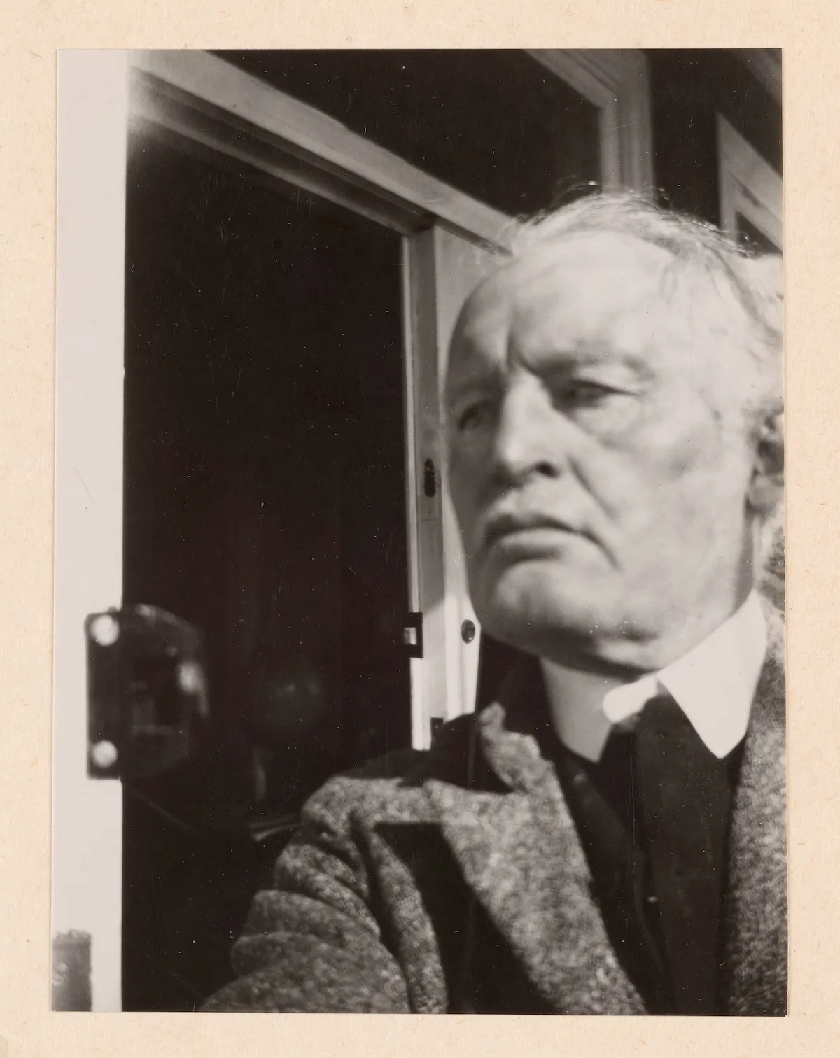 Siyah beyaz bu otoportrede, saçları dağılmış ve yüzünde ciddi bir ifade olan Edvard Munch, 1930'larda Ekely'deki evinde, pencere veya kapı pervazının önünde, beyaz yakalı gömlek ve yünlü bir ceketle sağa doğru hafifçe yukarı bakarken görülüyor.