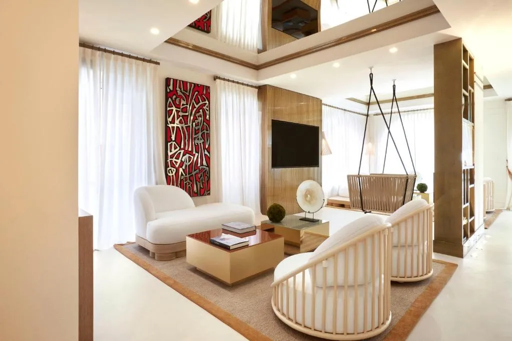 Lüks bir otel süitindeki modern yaşam alanı, beyaz uzanma koltuğu, iki ahşap döşemeli tekli koltuk, kırmızı soyut duvar sanatı, duvara monte televizyon ve asılı bir salıncak sandalye ile zarif ve aydınlık bir iç mekanı gösteriyor.