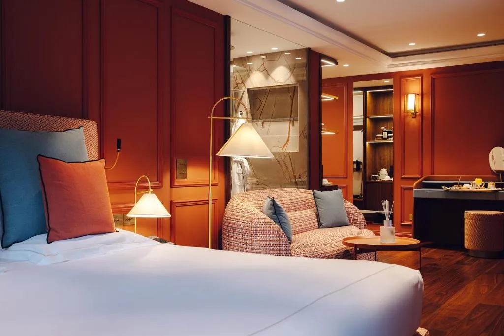 Terracota rengi panelli duvarlara sahip lüks bir otel odasında, beyaz çarşaflı konforlu bir yatak, desenli kumaş kaplı modern bir koltuk, zarif bir lambader, ahşap zemin ve şık makyaj masası ile sıcak aydınlatma davetkar bir atmosfer yaratıyor.