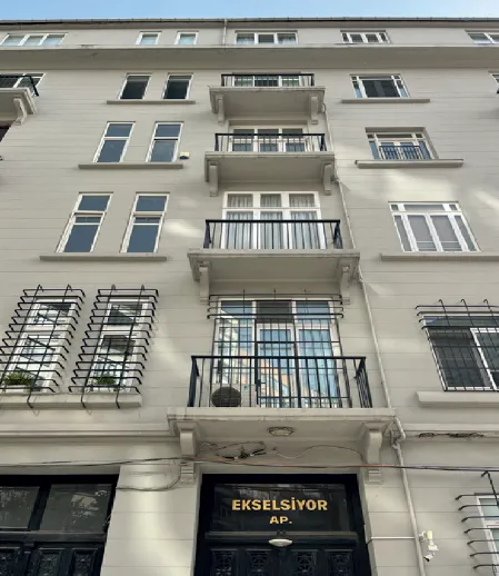 Ekselsiyor Apartmanı'nın alt kısmından yukarıya doğru çekilmiş bu detaylı fotoğrafta, açık gri renkli cephesi, her katında sıralanmış çeşitli pencereleri, siyah ferforje balkonları ve girişteki EKSELSİYOR AP. yazısı net bir şekilde görülmektedir.