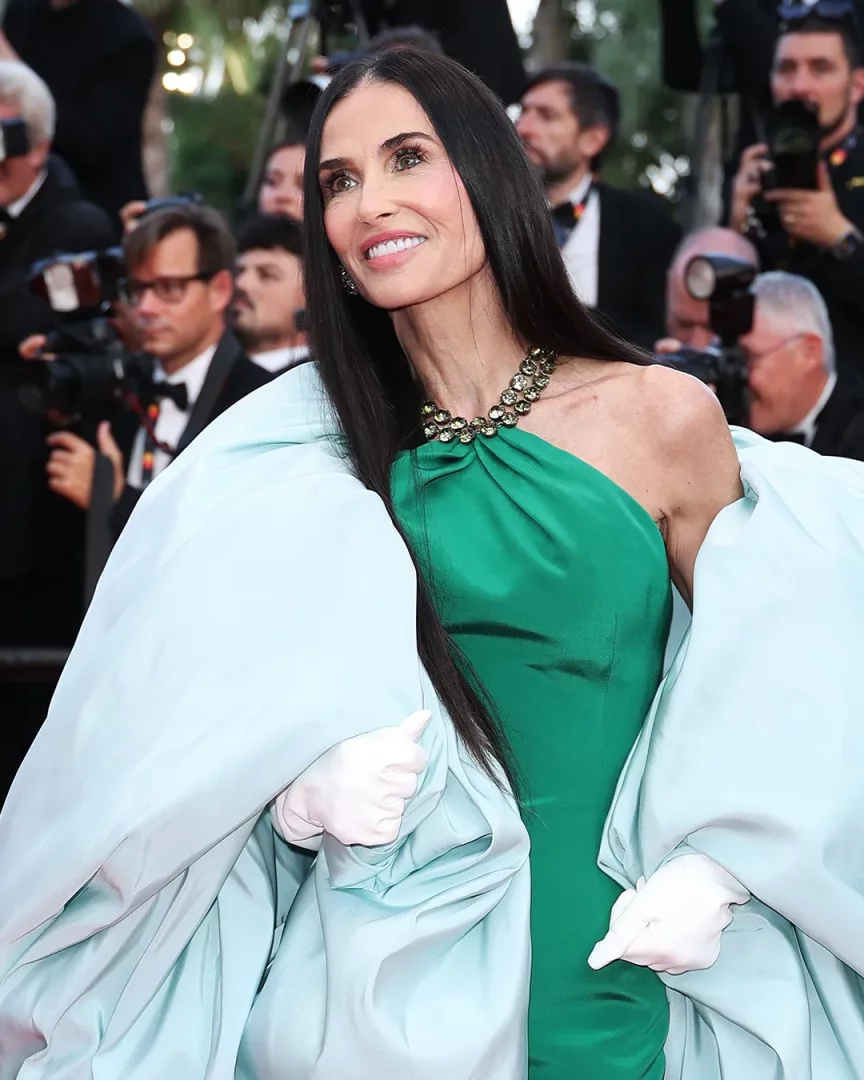 Demi Moore, 2025 Cannes Film Festivali'nde Chopard imzalı göz alıcı gerdanlığı, yeşil tek omuz elbisesi ve açık mavi, hacimli peleriniyle kırmızı halıda zarif bir gülümsemeyle poz veriyor, saçları omzuna dökülüyor.