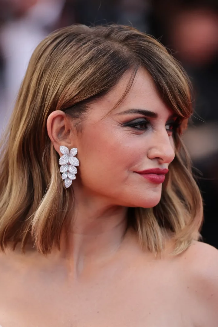 Penelope Cruz, Cannes Film Festivali kırmızı halısında yandan çekilen yakın planda, dalgalı kahverengi saçları, kırmızı ruju ve belirgin göz makyajıyla zarif görünürken, kulaklarındaki yaprak formunda, pırlantalarla bezeli ışıltılı Pasquale Bruni mücevherleri ile dikkat çekiyor.