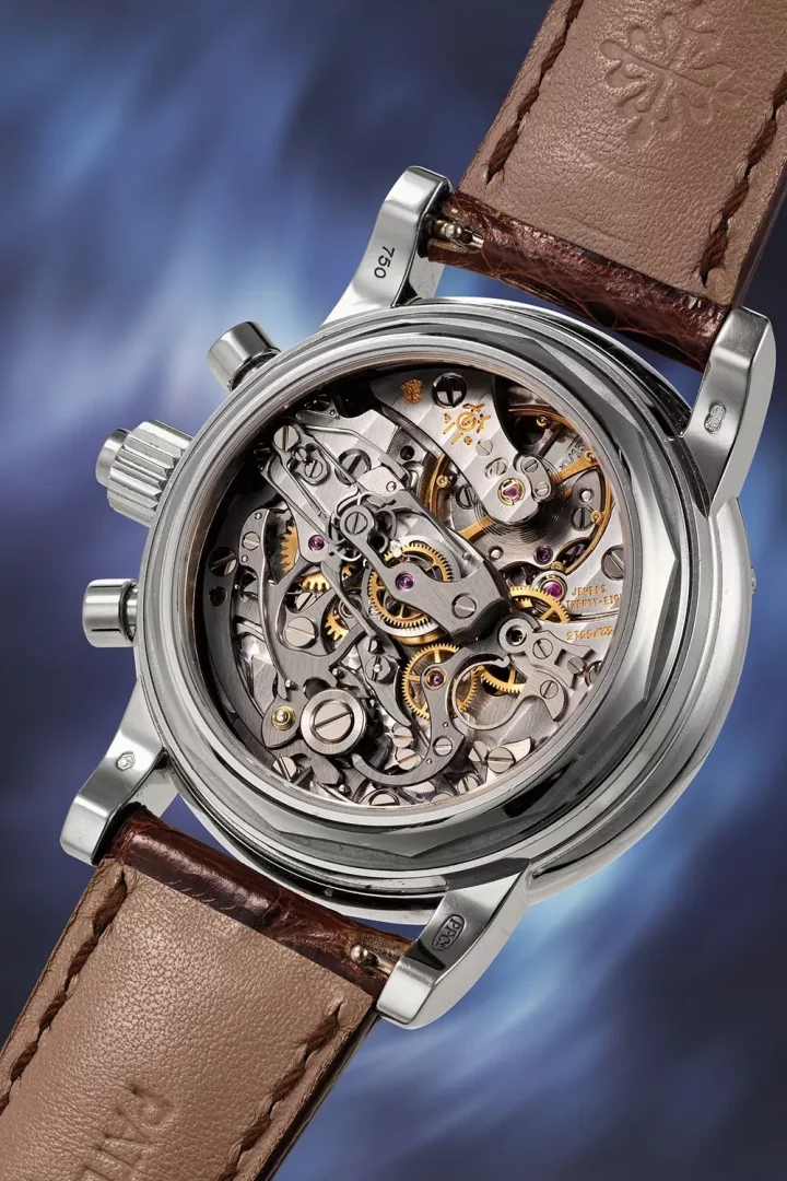 Şeffaf arka kapağı sayesinde gümüş renkli kasası ve kahverengi timsah derisi kayışıyla görülen, Patek Philippe mekanik saatinin karmaşık çarklar, köprüler ve yakut taşlarıyla bezeli hassas iç işleyişini yakından gösteren, ustalıkla işlenmiş detaylı bir görüntü.