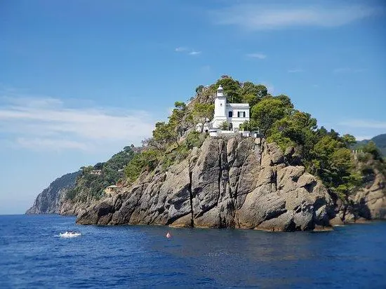 Güneşli bir havada, masmavi sularla çevrili, yeşil ağaçlar ve kayalıklarla kaplı bir burun üzerinde yükselen beyaz Portofino Feneri ve yanındaki binası, uzakta yüzen küçük bir tekne ve kırmızı bir şamandıra ile sakin deniz manzarası.
