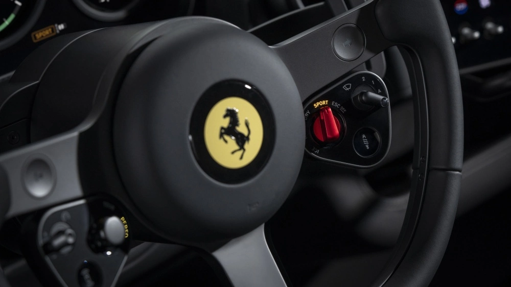 Bir Ferrari direksiyonunun yakın çekiminde, ortadaki sarı zeminli siyah şaha kalkan at logosu ve sağda kırmızı renkte aydınlatılmış SPORT mod tuşu ile silecek ve far kontrolleri gibi alüminyum detaylı fonksiyonel düğmeler belirgin şekilde görülüyor.