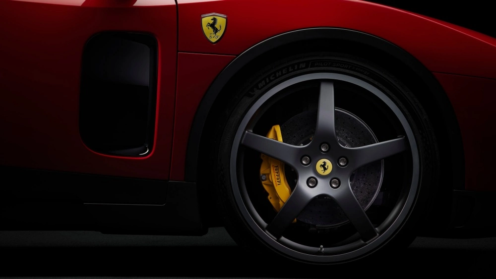 Koyu kırmızı bir Ferrari'nin ön tarafının yakın çekiminde, Ferrari logosu ve Michelin Pilot Sport yazısı görünen mat siyah beş kollu jant tasarımı ile sarı renkli Ferrari yazılı kaliperlere sahip, gelişmiş karbon seramik fren sistemi dikkat çekiyor.