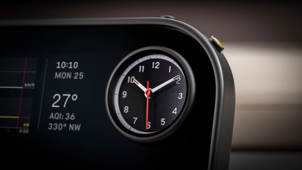 Bir Ferrari'nin modern iç mekanında, merkez konsol üzerine entegre edilmiş parlak OLED ekranın solunda 10:10 saati, pazartesi 25 tarihi ve 27°C sıcaklık bilgileri gösterilirken, sağ tarafında kırmızı saniye ibresi ve beyaz rakamlarla şık bir analog saat yer alıyor.