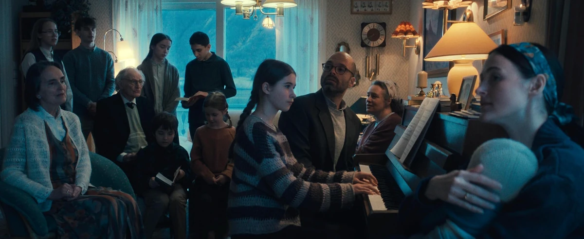 Fjord filmi aile sahnesinde, loş ışıklı bir oturma odasında piyano çalan genç bir kızın etrafında, bebekten yaşlıya kadar farklı kuşaklardan birçok aile üyesi toplanmış, pencereden görünen dağ manzarası eşliğinde müzik dinliyor gibi görünüyor.
