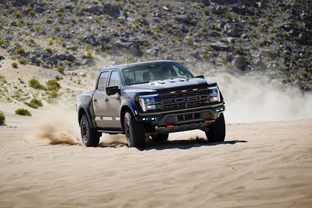 Sert offroad koşulları için tasarlanmış, kumlu bir çölde tekerleklerinden yoğun tozlar kaldırarak hızla ilerleyen koyu renkli bir Ford F-150 Raptor pick-up aracı, gücünü ve arazi yeteneğini sergiliyor.