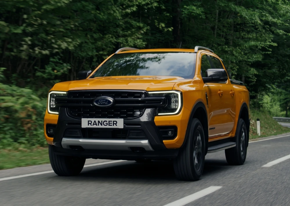 Canlı sarı rengiyle yeni nesil Ford Ranger pickup, yemyeşil ağaçların çevrelediği asfalt bir yolda önden hafif açıyla ilerlerken, gündüz farları yanık ve güçlü arazi yeteneklerini ima eden kararlı bir duruş sergiliyor.