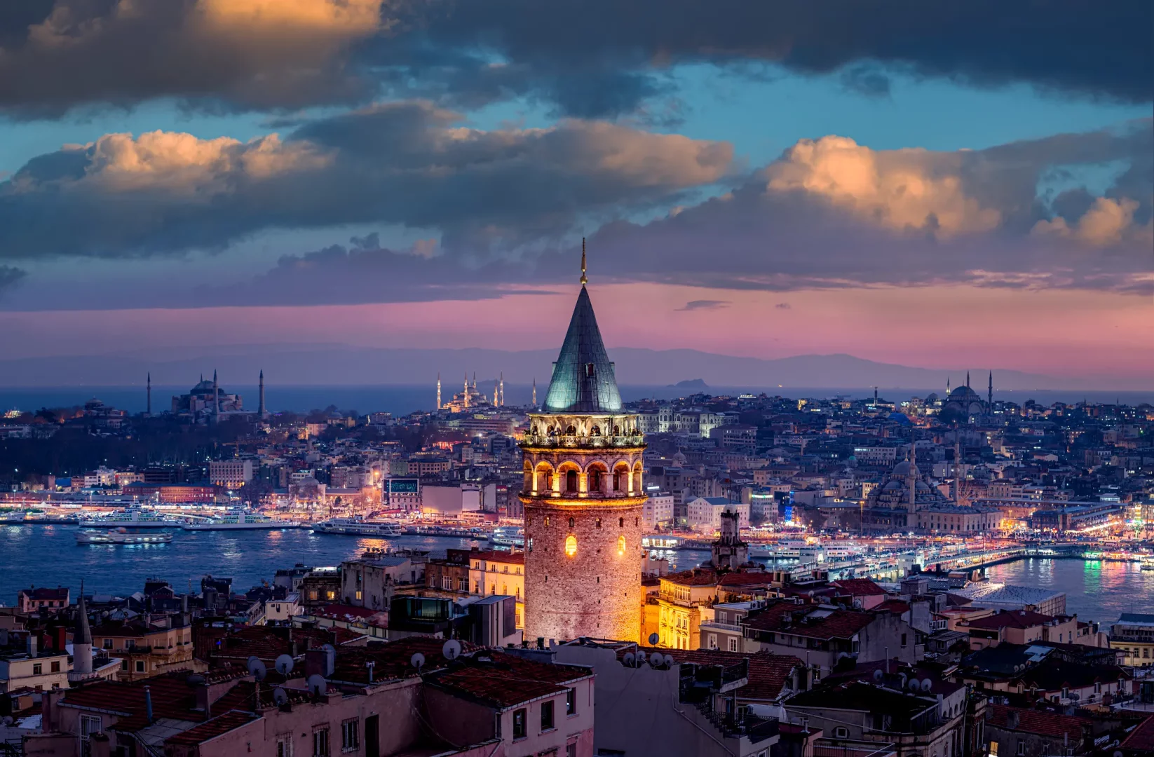 Galata Kulesi'nin İstanbul'un eşsiz gün batımı manzarasıyla parladığı, Haliç'teki gemilerin ışıklarıyla birleştiği ve Ayasofya ile Sultanahmet Camii gibi tarihi yapıların silüetlerinin de göründüğü nefes kesici bir şehir panoraması.