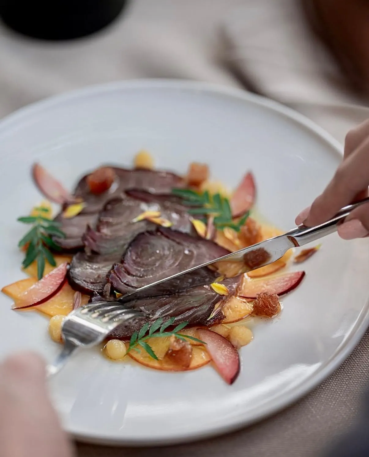 Gia Urla restoranında sunulan gurme bir tabakta, ince dilimlenmiş koyu renkli etin veya sebzenin, taze meyve dilimleri, nohut ve yeşilliklerle zenginleştirilmiş sunumunu gümüş çatal bıçakla kesen bir el, Urla lezzetlerini deneyimlerken estetik bir görsel şölen sunuyor.