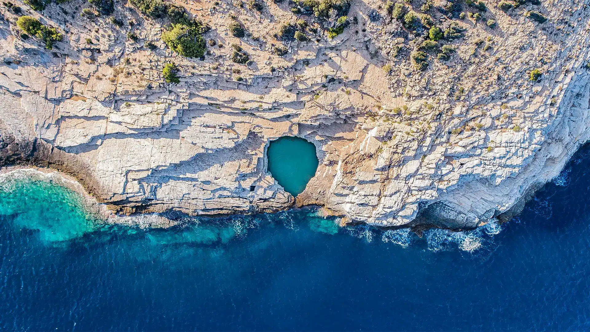 Giola'nın güneşli engebeli kayalık kıyı şeridinin kuşbakışı görünümünde, berrak turkuaz suya sahip göz alıcı doğal havuz ve kıyıyı saran koyu mavi Ege Denizi'nin büyüleyici manzarası belirgin bir şekilde sunuluyor.
